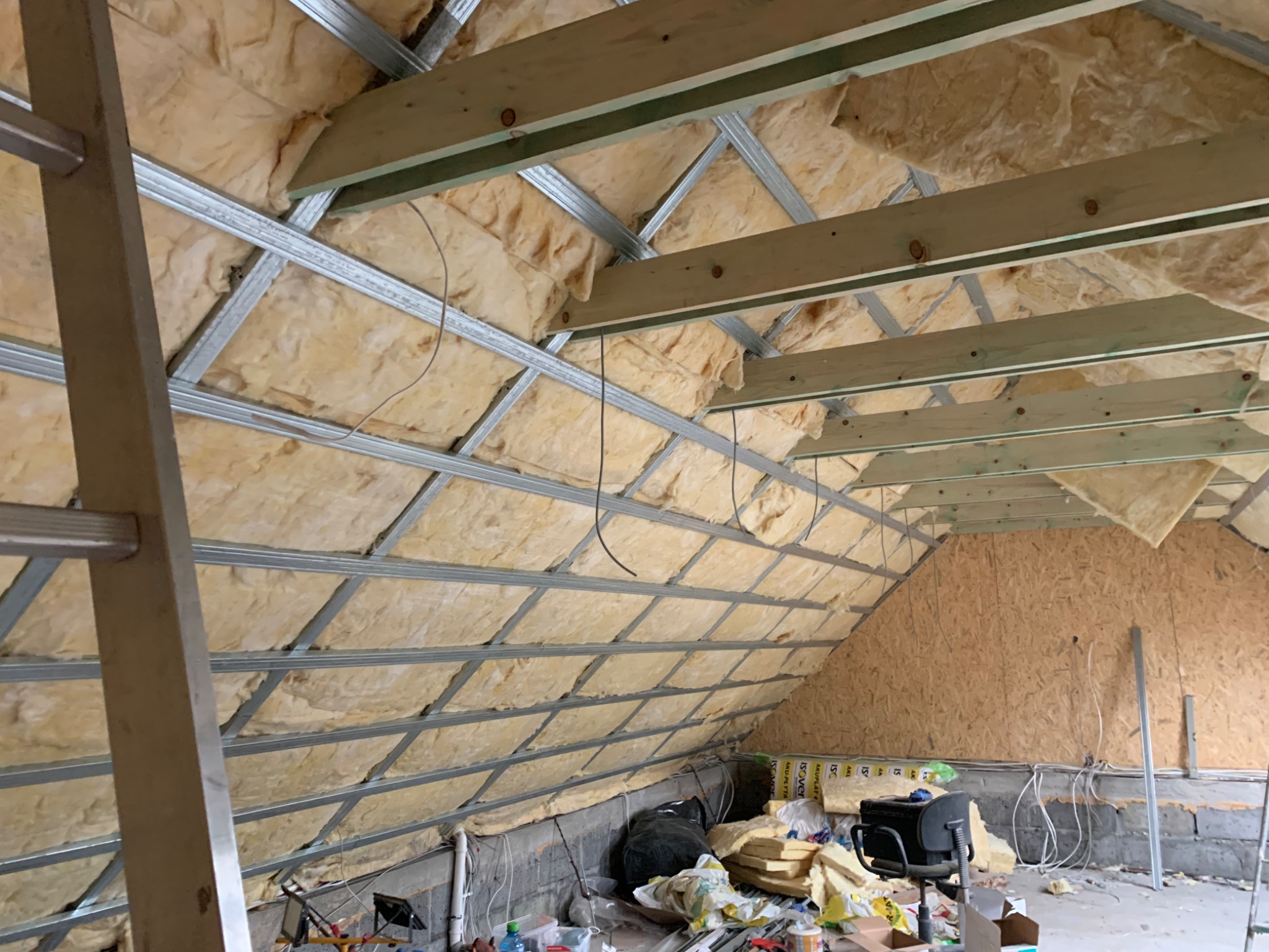 Izolacja poddasza wełną mineralną, widoczne metalowe profile konstrukcyjne i drewniane belki, częściowo wykończona ściana z płyty OSB, bałagan na podłodze.