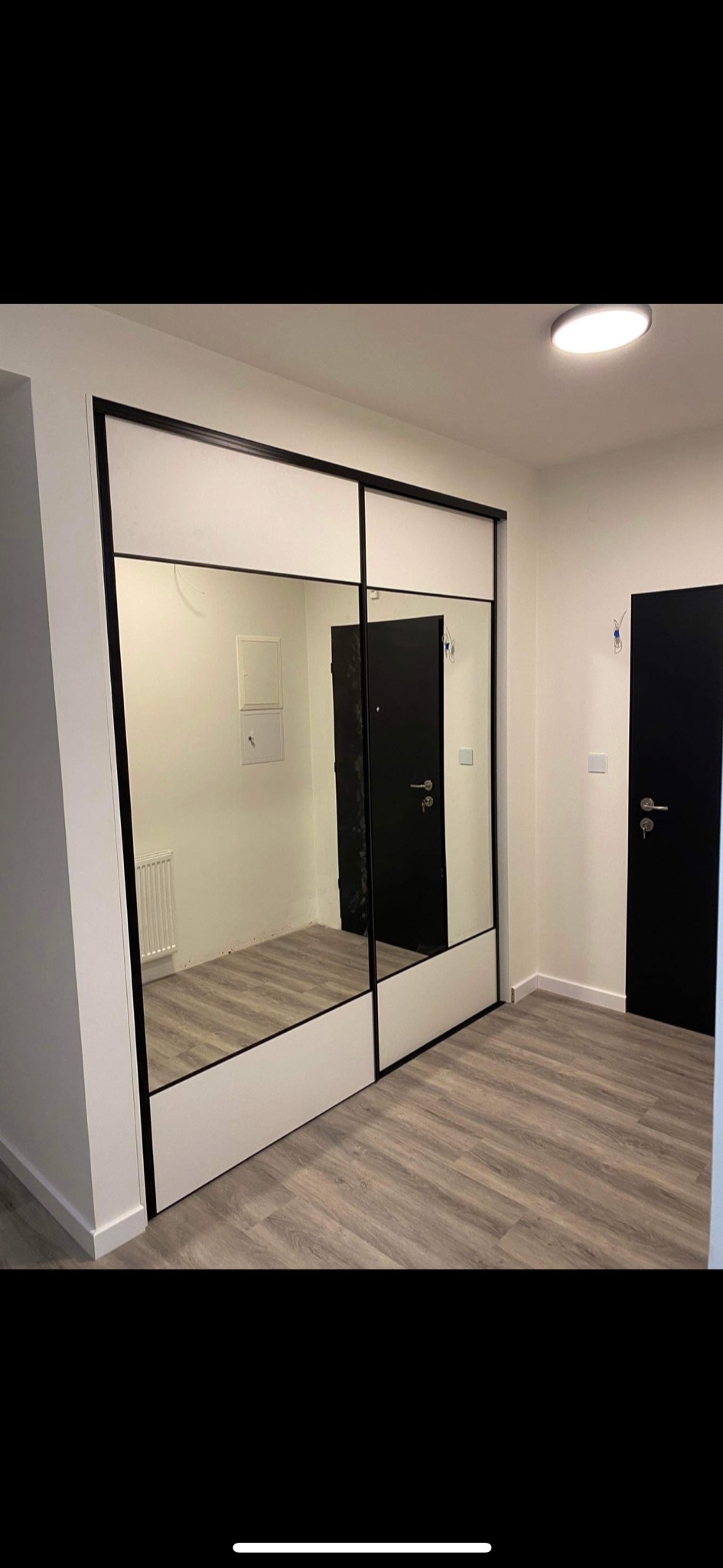 Nowoczesna szafa wnękowa z przesuwnymi drzwiami lustrzanymi, czarne ramy, białe panele górne, parkiet w odcieniach szarości, Kraków.