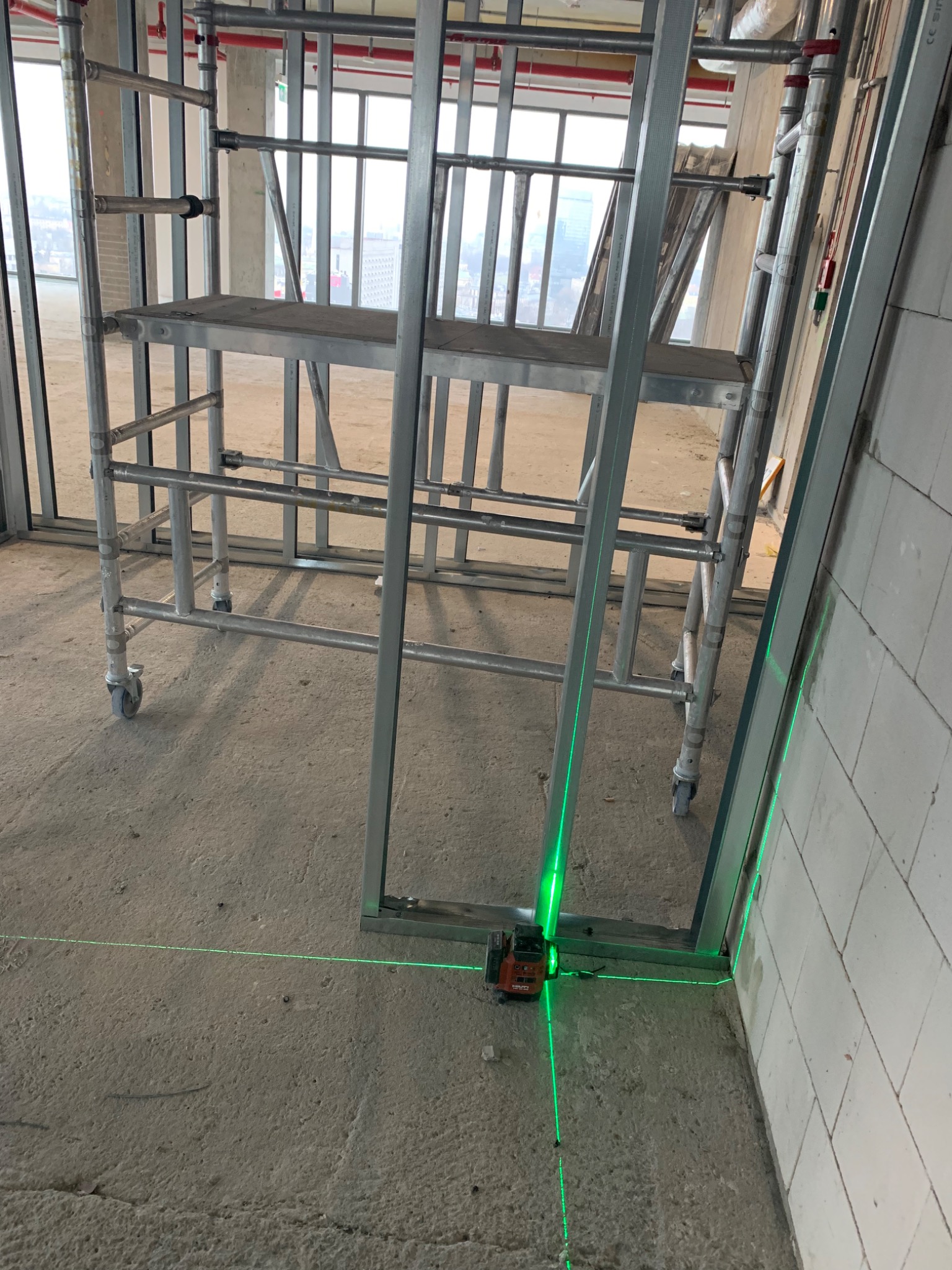 Ustawianie metalowej konstrukcji działowej za pomocą lasera krzyżowego, widoczne rusztowanie i surowy stan pomieszczenia.