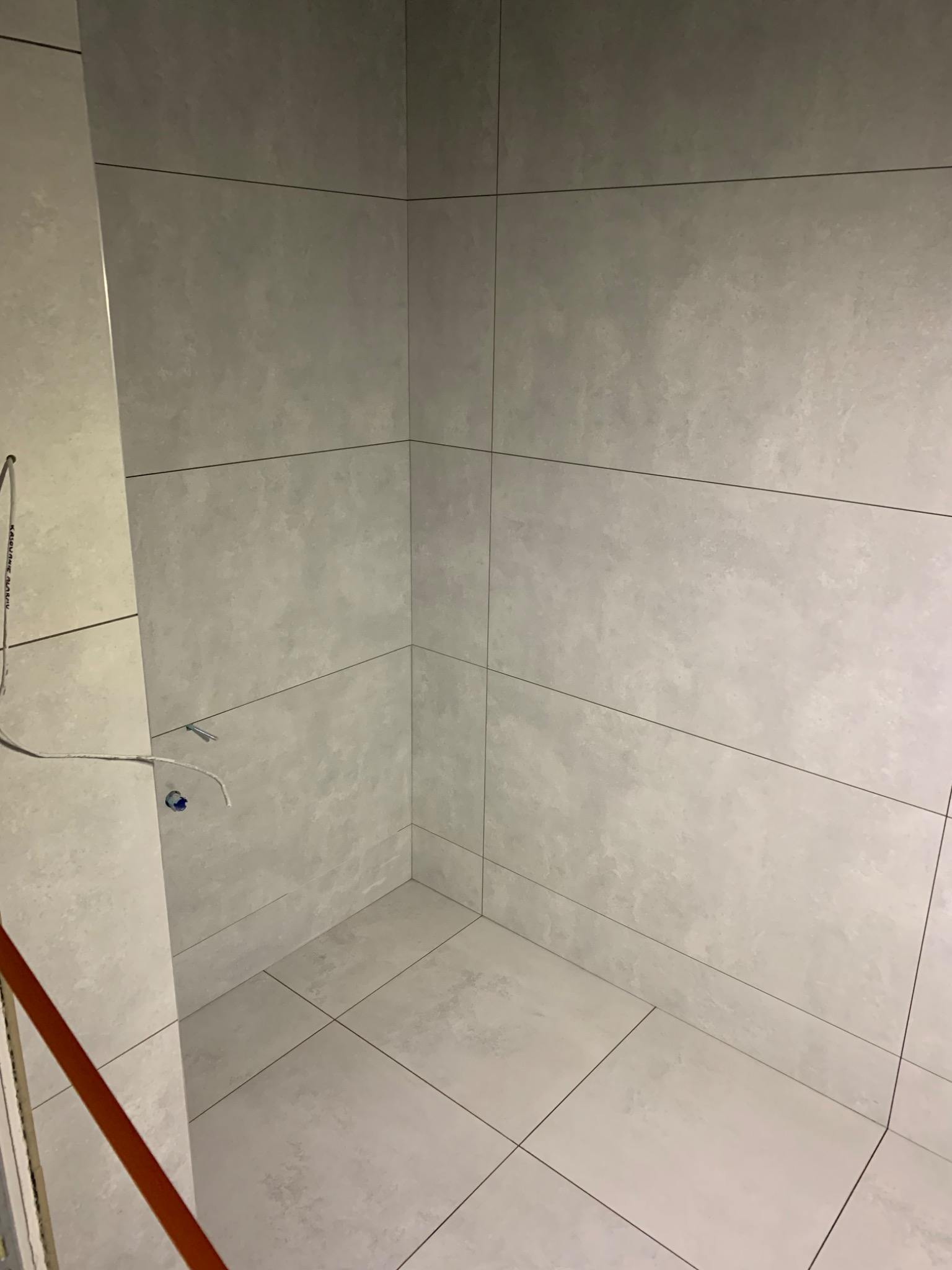 Wykończona kabina prysznicowa z szarymi, matowymi płytkami o dużym formacie, widoczne wystające kable i rurki przed montażem armatury.