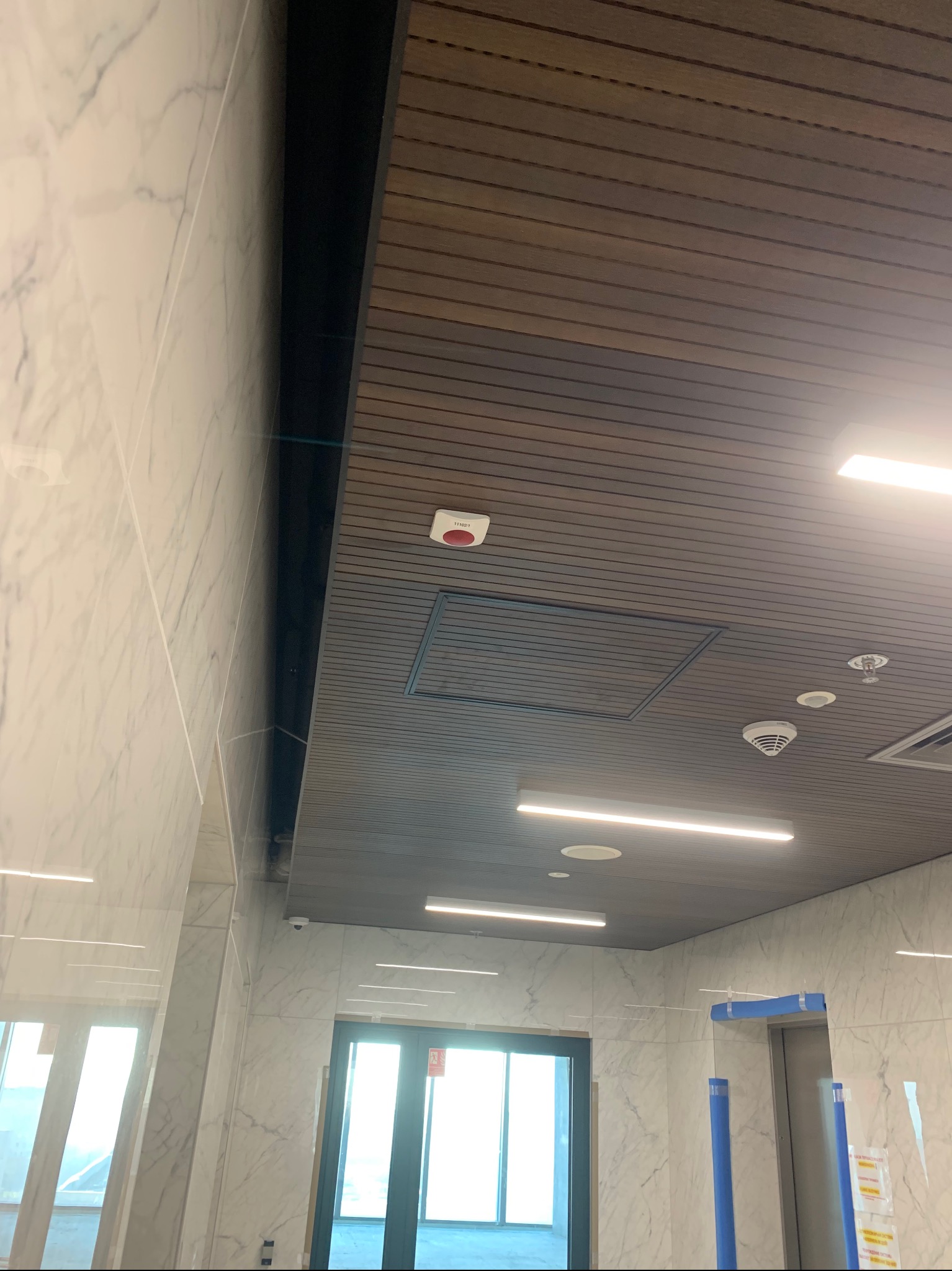Detale wykończenia wnętrza: drewnopodobny sufit z widocznymi elementami instalacji, marmurowe ściany i wejście do windy.