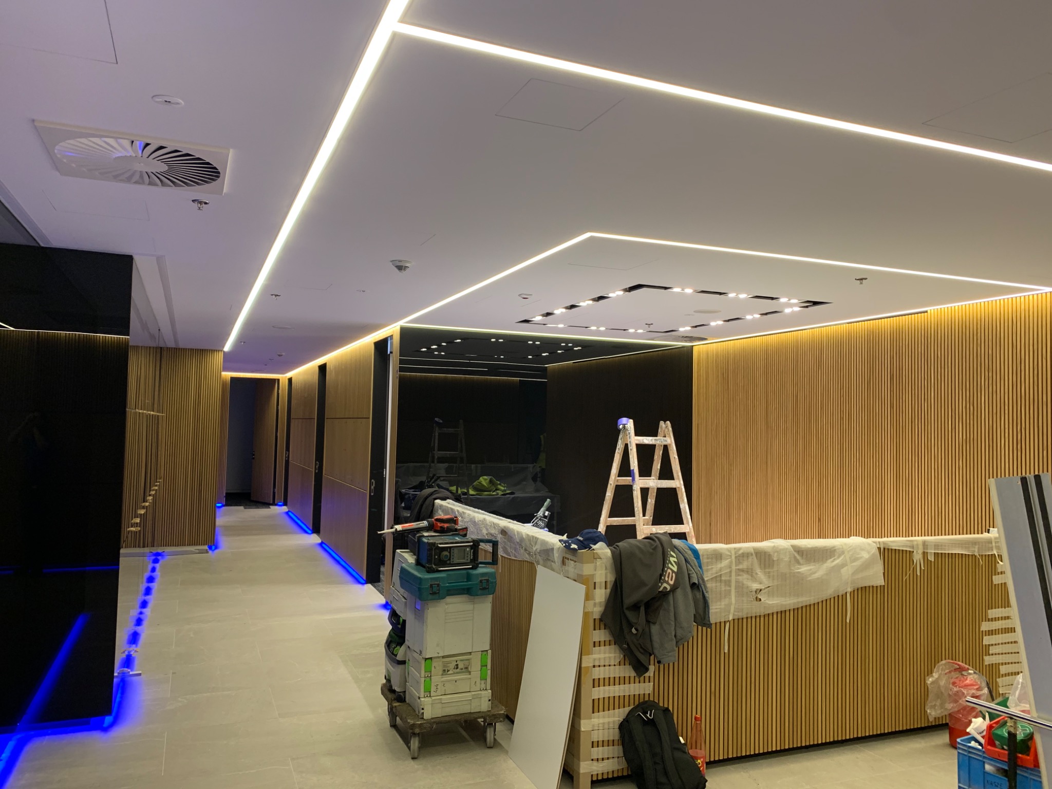 Wnętrze biura w trakcie prac wykończeniowych: widoczne drewniane panele ścienne, oświetlenie liniowe LED na suficie, narzędzia i drabina, niebieskie oświetlenie podłogowe.