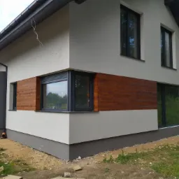 Nowoczesny dom z jasną elewacją, drewnianą okładziną pod oknami i ciemną podmurówką, widok z boku w trakcie prac wykończeniowych.