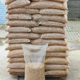 Wysoki stos worków z pelletem na palecie, jeden worek stoi przed stosem, widoczne tło betonowe i fragment zieleni.