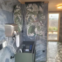 Nowoczesna łazienka z umywalką w kolorze oliwkowym, suszarką do rąk i płytkami imitującymi marmur w odcieniach szarości i błękitu, akcentem jest tapeta z motywem tropikalnym i ptakiem.