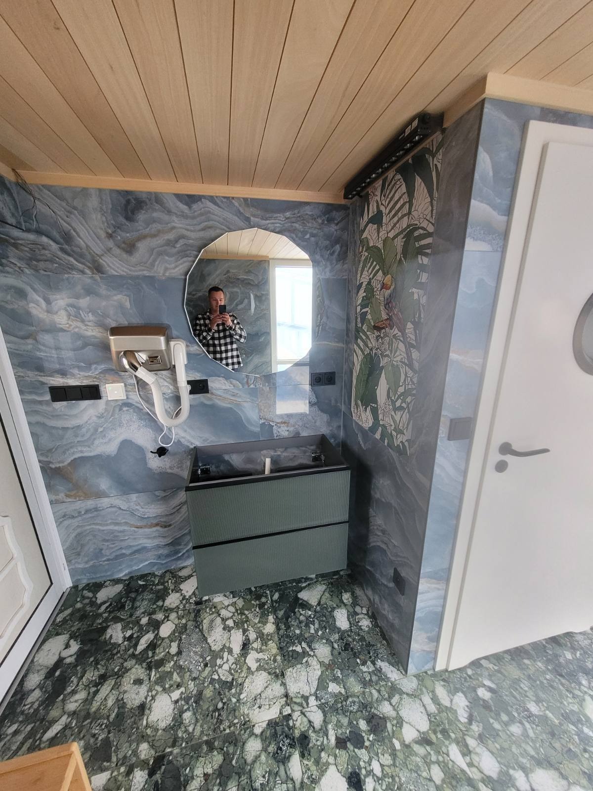 Ukończona łazienka z marmurową podłogą i ścianami w odcieniach niebieskiego, drewniany sufit, okrągłe lustro odbijające fotografa, nowoczesna szafka pod umywalkę i suszarka do rąk.