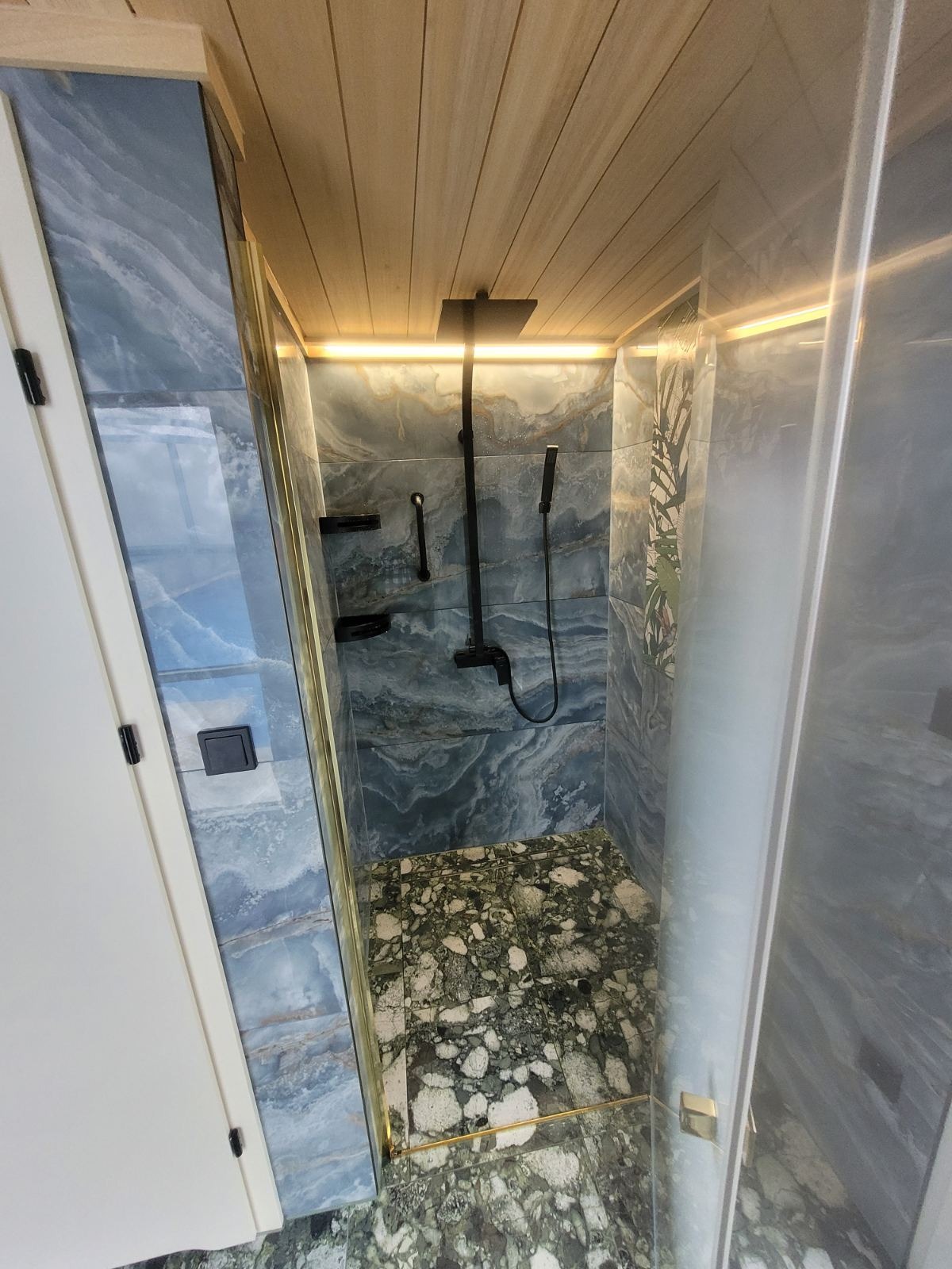 Nowoczesna kabina prysznicowa z niebieskimi płytkami imitującymi marmur, drewnianym sufitem z oświetleniem LED, czarną armaturą i podłogą z zielonego kamienia.