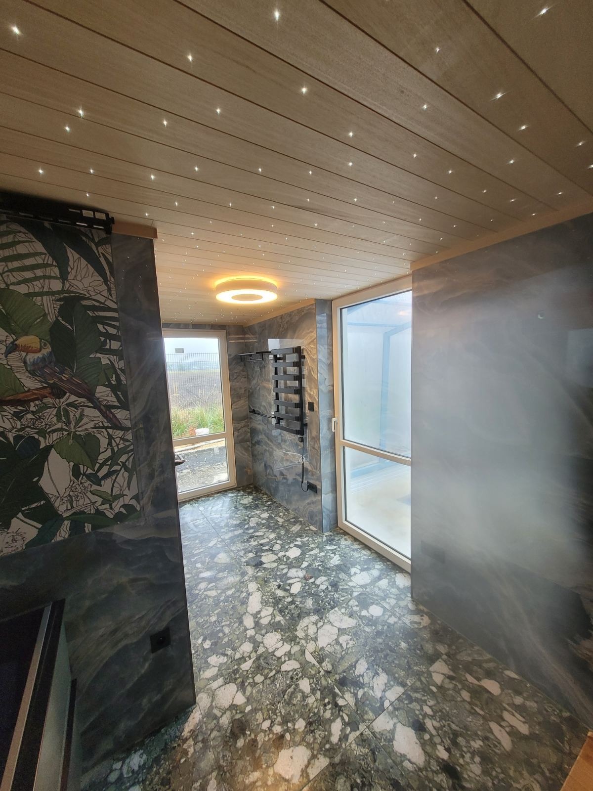 Nowoczesna łazienka z marmurową podłogą i ścianami w odcieniach szarości, drewnianym sufitem z punktowym oświetleniem LED oraz okrągłym, podwieszanym światłem, grzejnik drabinkowy na ścianie, drzwi...