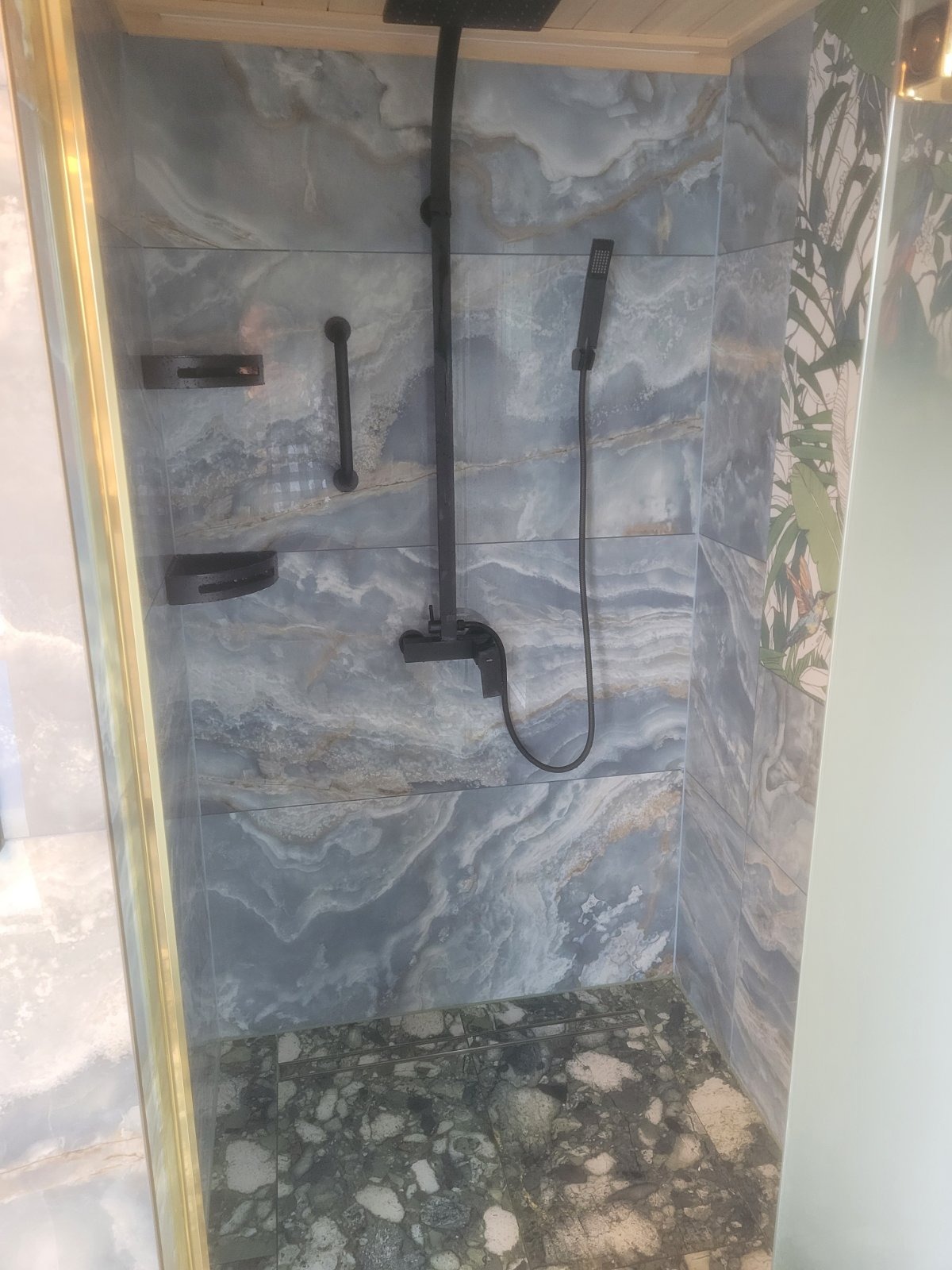 Kabina prysznicowa z niebiesko-szarymi płytkami imitującymi marmur, czarną armaturą, półkami i uchwytem, oraz podłogą z ciemnego lastryko. Na jednej ze ścian widoczny wzór z motywem roślinnym.