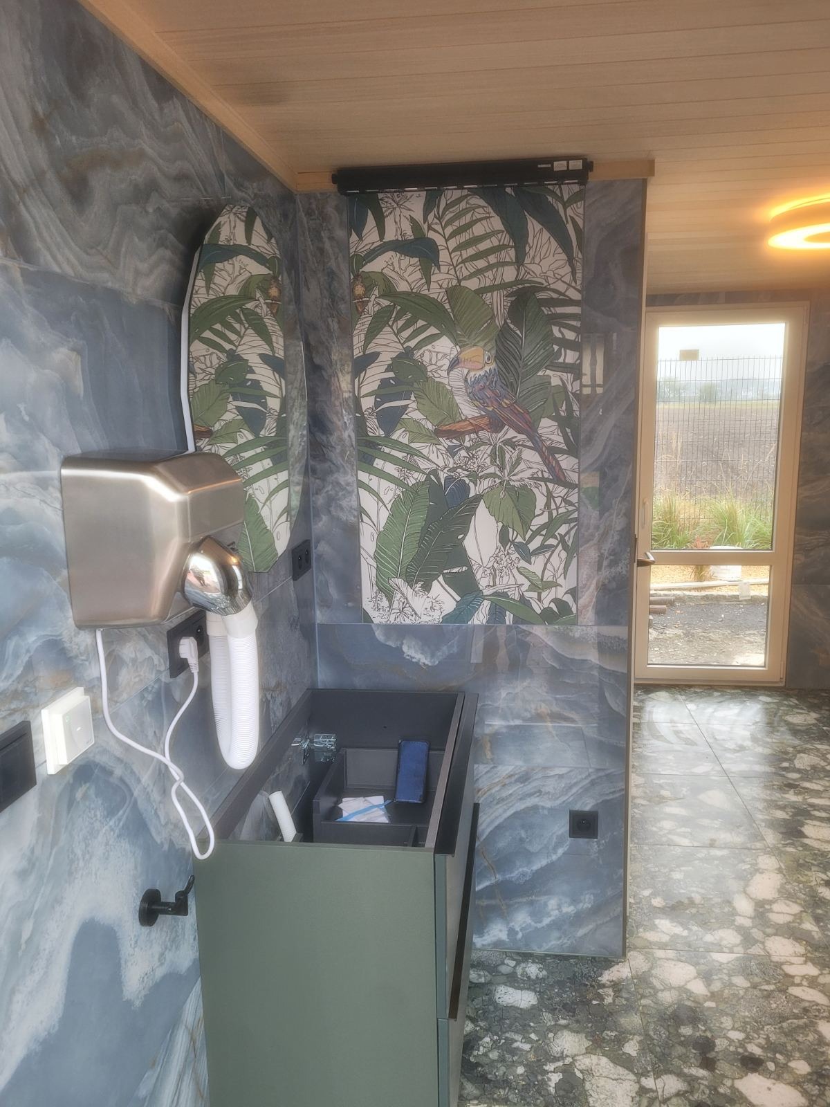 Nowoczesna łazienka z umywalką w kolorze oliwkowym, suszarką do rąk i płytkami imitującymi marmur w odcieniach szarości i błękitu, akcentem jest tapeta z motywem tropikalnym i ptakiem.