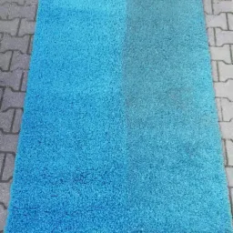 Turkusowy dywan shaggy, po lewej stronie świeżo wyczyszczony, po prawej widoczna różnica przed czyszczeniem, ułożony na szarej kostce brukowej.