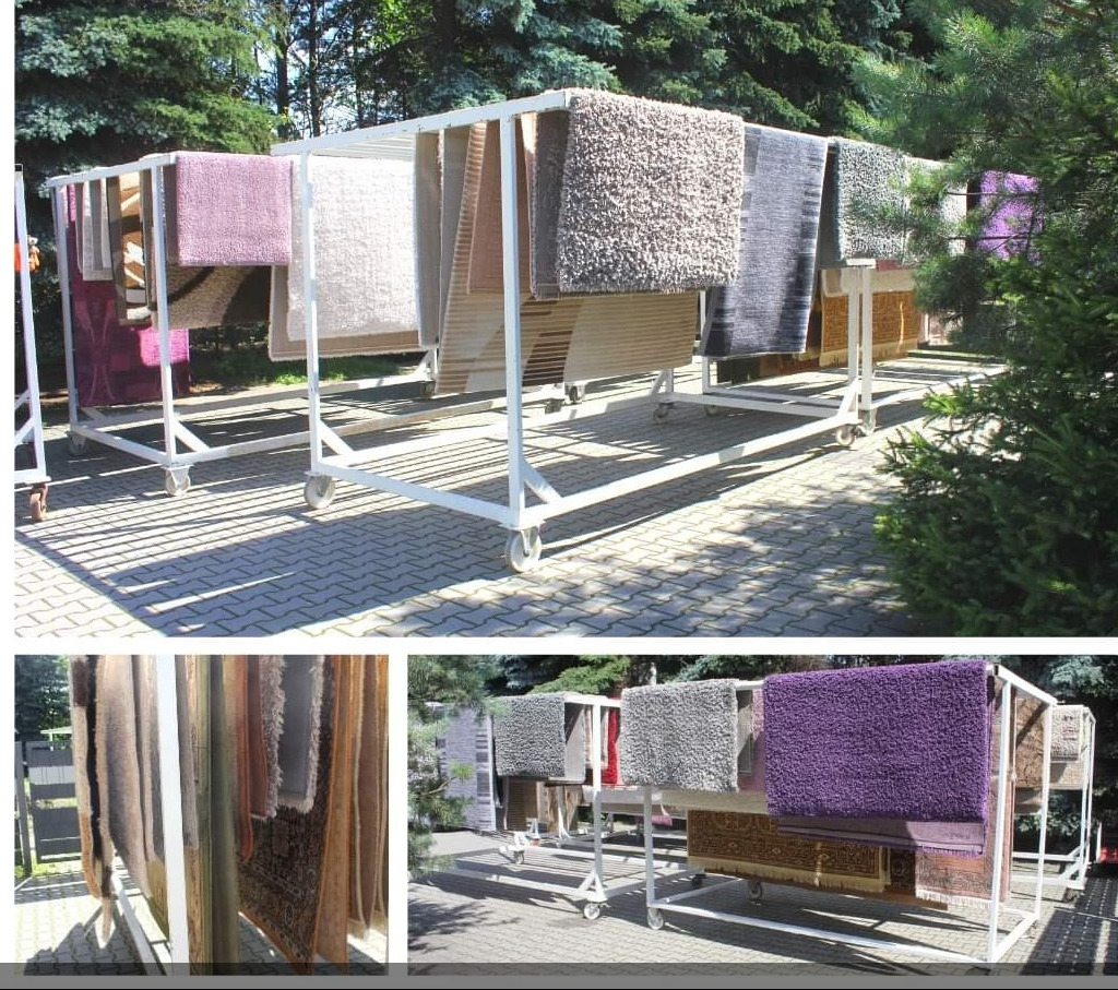 Różnokolorowe dywany suszone na metalowych stojakach z kółkami na zewnątrz, na brukowanym podłożu, w tle drzewa.