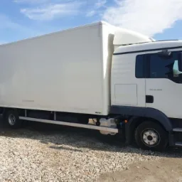 Biały samochód dostawczy MAN TGL 12.250 z zabudową kontenerową, zaparkowany na żwirowym podłożu w słoneczny dzień.