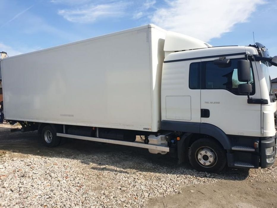 Biały samochód dostawczy MAN TGL 12.250 z zabudową kontenerową, zaparkowany na żwirowym podłożu w słoneczny dzień.