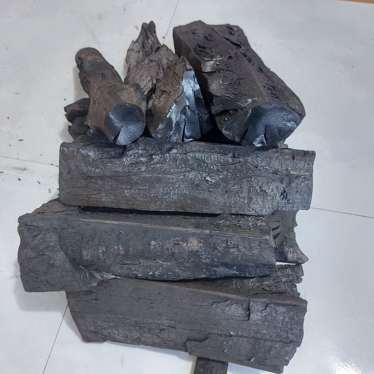 Stos ułożonych kawałków brykietu drzewnego o nieregularnych kształtach, widoczne ślady obróbki i naturalna struktura drewna, na jasnym tle.