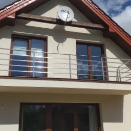 Elewacja domu z balkonem zabezpieczonym prostą, poziomą konstrukcją ze stali nierdzewnej; w oknach odbijają się chmury.