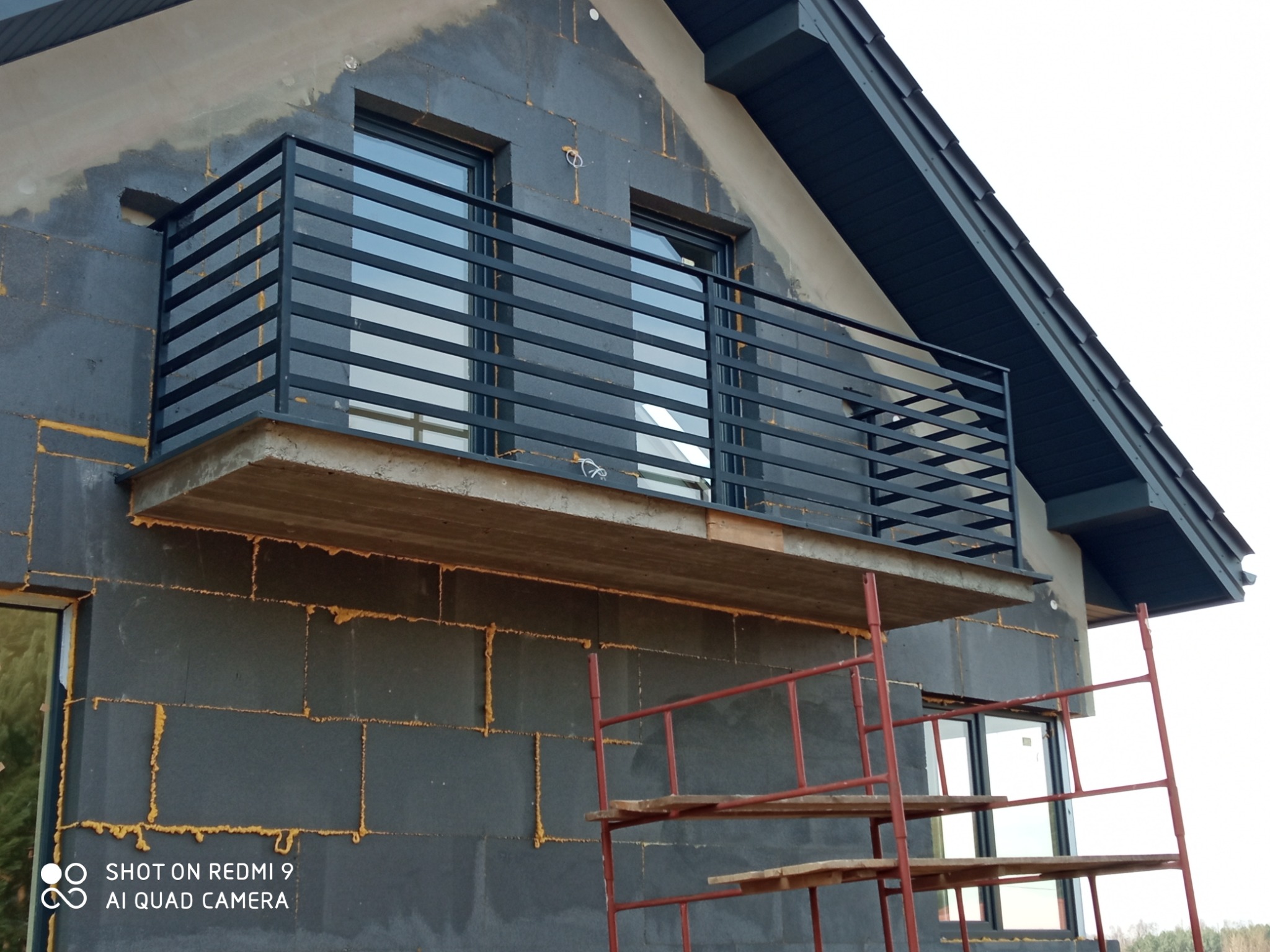 Surowy balkon z czarną, poziomą balustradą na tle szarej, nieotynkowanej ściany domu w trakcie budowy, widoczne rusztowanie.