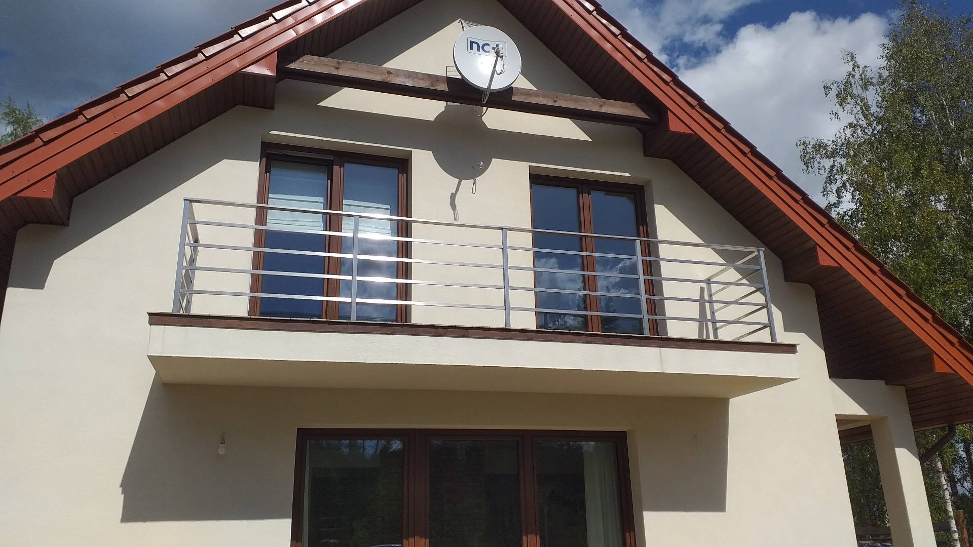 Elewacja domu z balkonem zabezpieczonym prostą, poziomą konstrukcją ze stali nierdzewnej; w oknach odbijają się chmury.