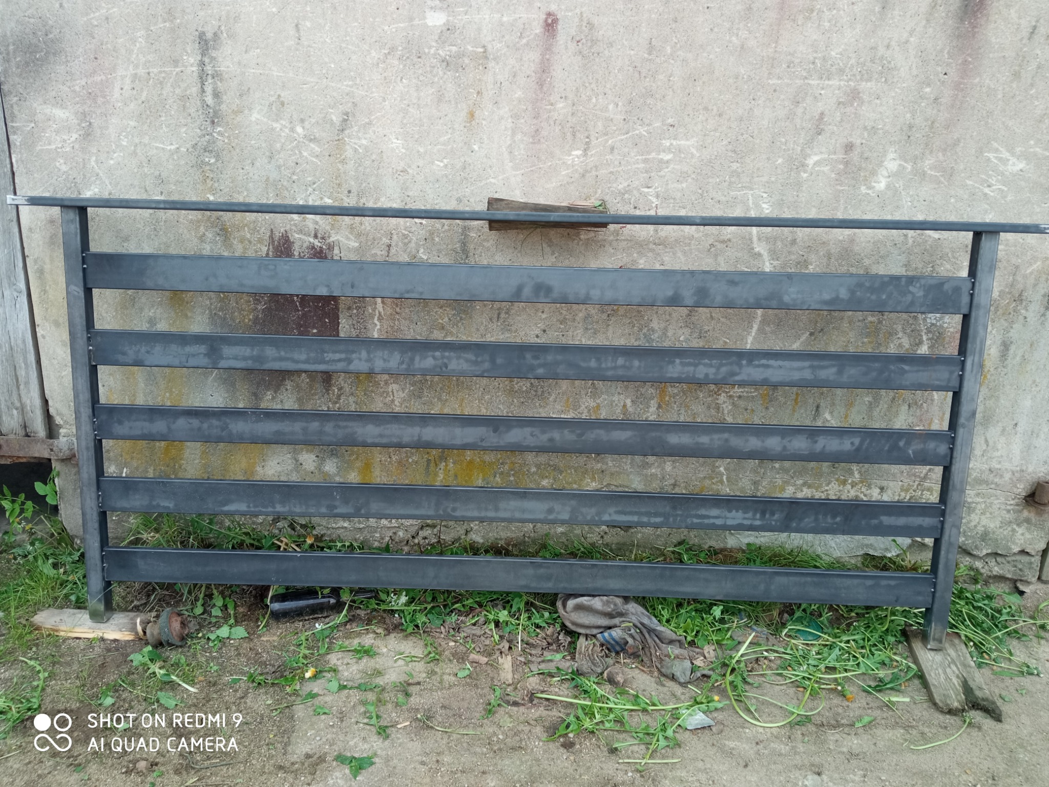 Surowa, spawana konstrukcja z metalowych profili, prawdopodobnie fragment przyszłego ogrodzenia, leżąca na ziemi przed betonową ścianą.