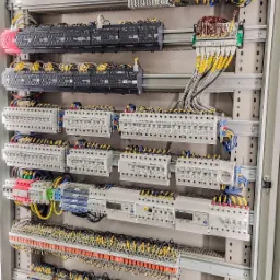 Rozdzielnica elektryczna z licznymi zabezpieczeniami, modułami i okablowaniem, widoczne bezpieczniki, przekaźniki i zasilacze, uporządkowane przewody z oznaczeniami.