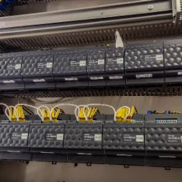 Panel sterowania z modułami Grenton, widoczne moduły I/O, moduły przekaźnikowe, moduły roletowe, okablowanie w żółtych i białych peszlach, całość zamontowana w szafie sterowniczej.