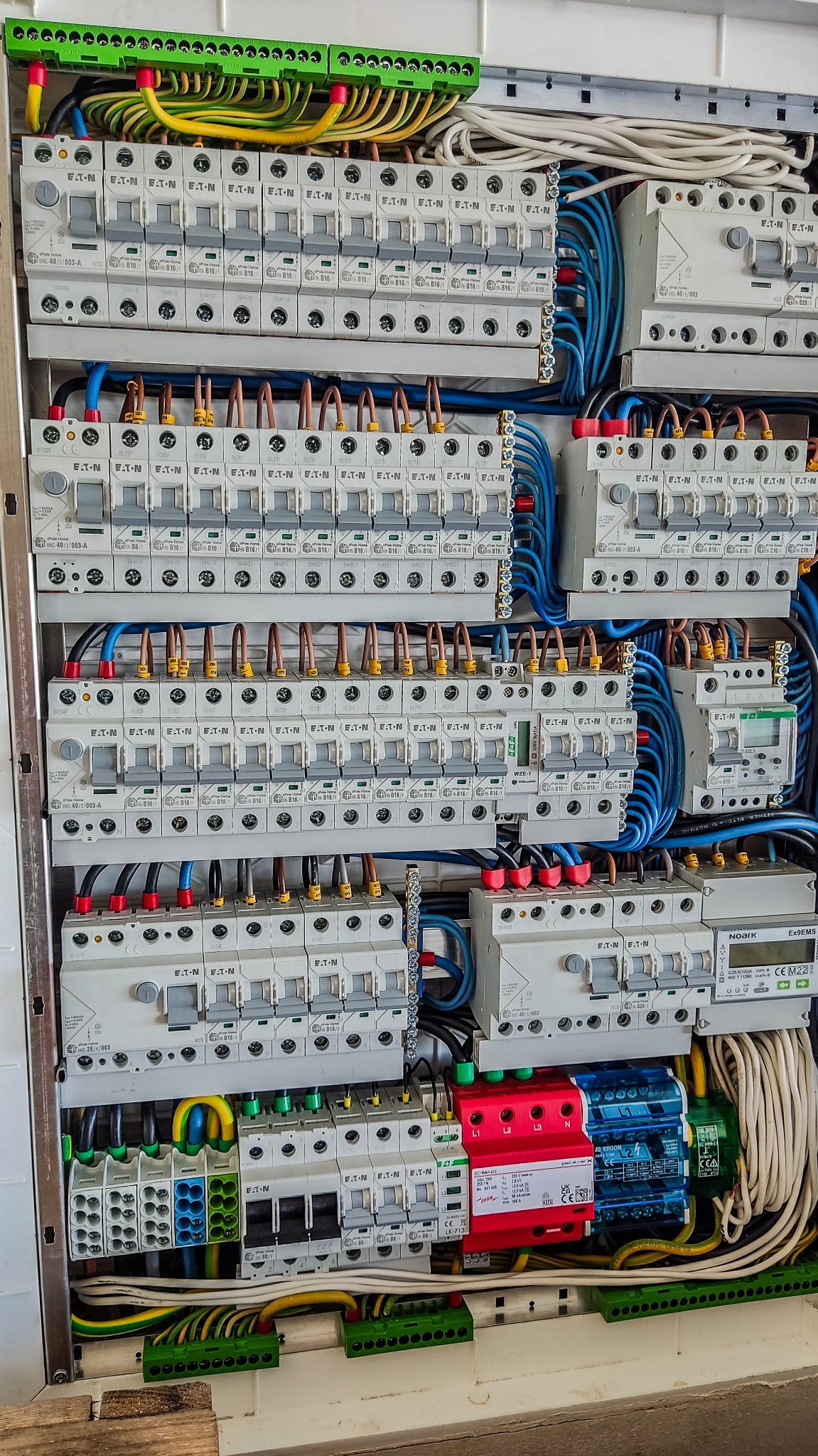 Szczegółowe ujęcie rozdzielnicy elektrycznej z licznymi bezpiecznikami Eaton, przewodami w kolorach żółto-zielonym, niebieskim i brązowym, oraz zabezpieczeniami przeciwprzepięciowymi Erom i Noark...