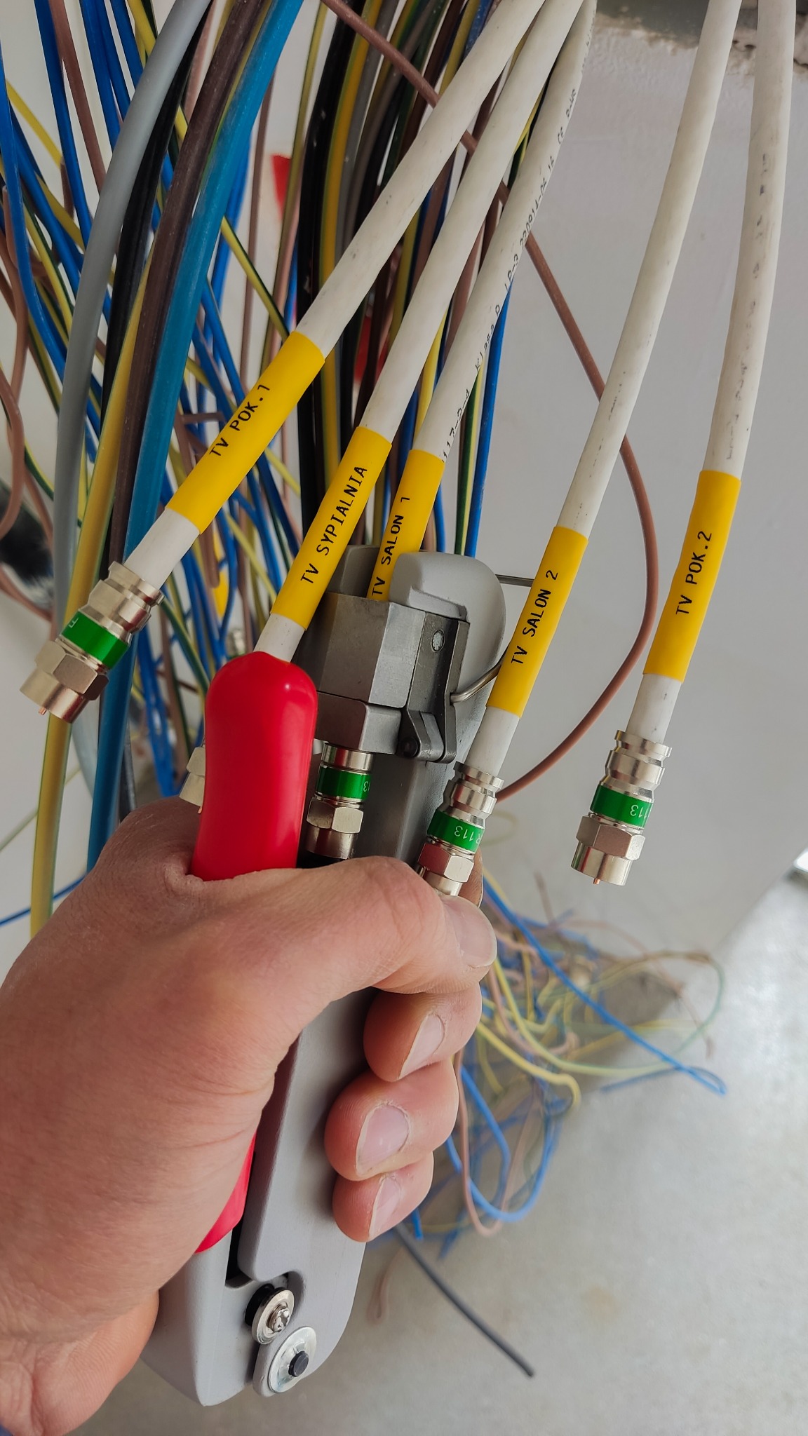 Ręka zaciska złączki F na kablach koncentrycznych, oznaczonych żółtymi etykietami z nazwami pomieszczeń; w tle wiązka kolorowych przewodów.