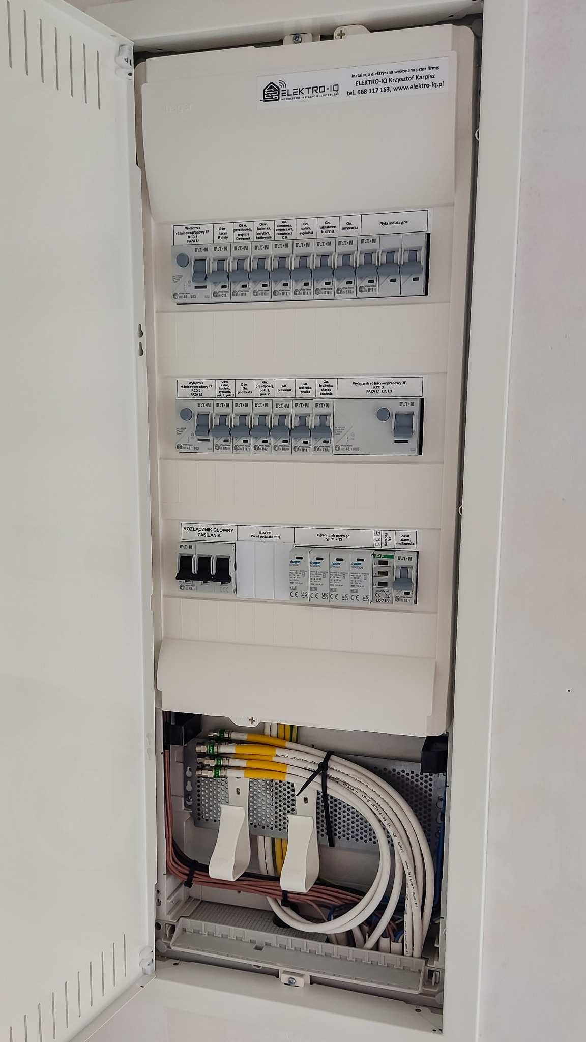 Otwarta skrzynka rozdzielcza z widocznymi bezpiecznikami, wyłącznikami różnicowoprądowymi, ogranicznikiem przepięć oraz uporządkowanymi przewodami elektrycznymi i kablami antenowymi. Widoczna...