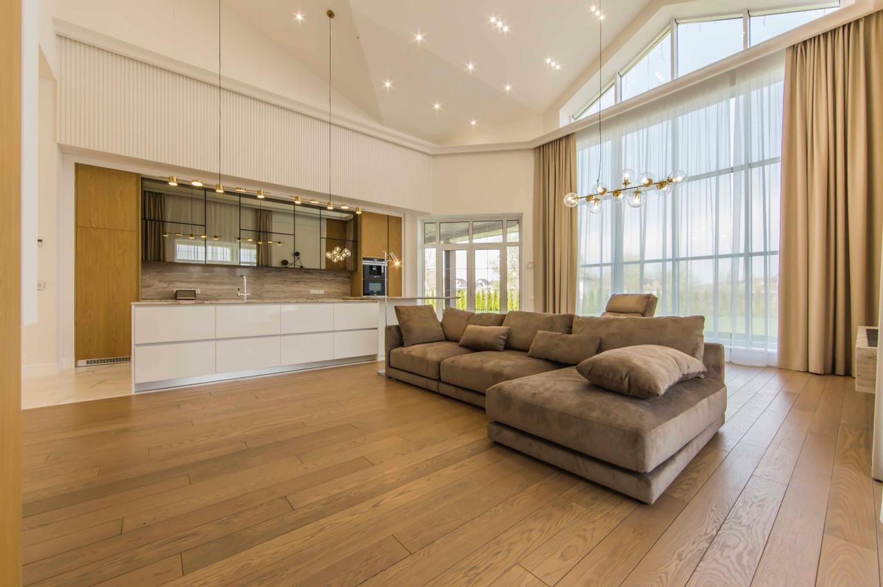 Przestronne, jasne wnętrze z dużym narożnikiem w odcieniach brązu, drewnianą podłogą i nowoczesną kuchnią w tle, oświetlone przez wysokie okna z beżowymi zasłonami.