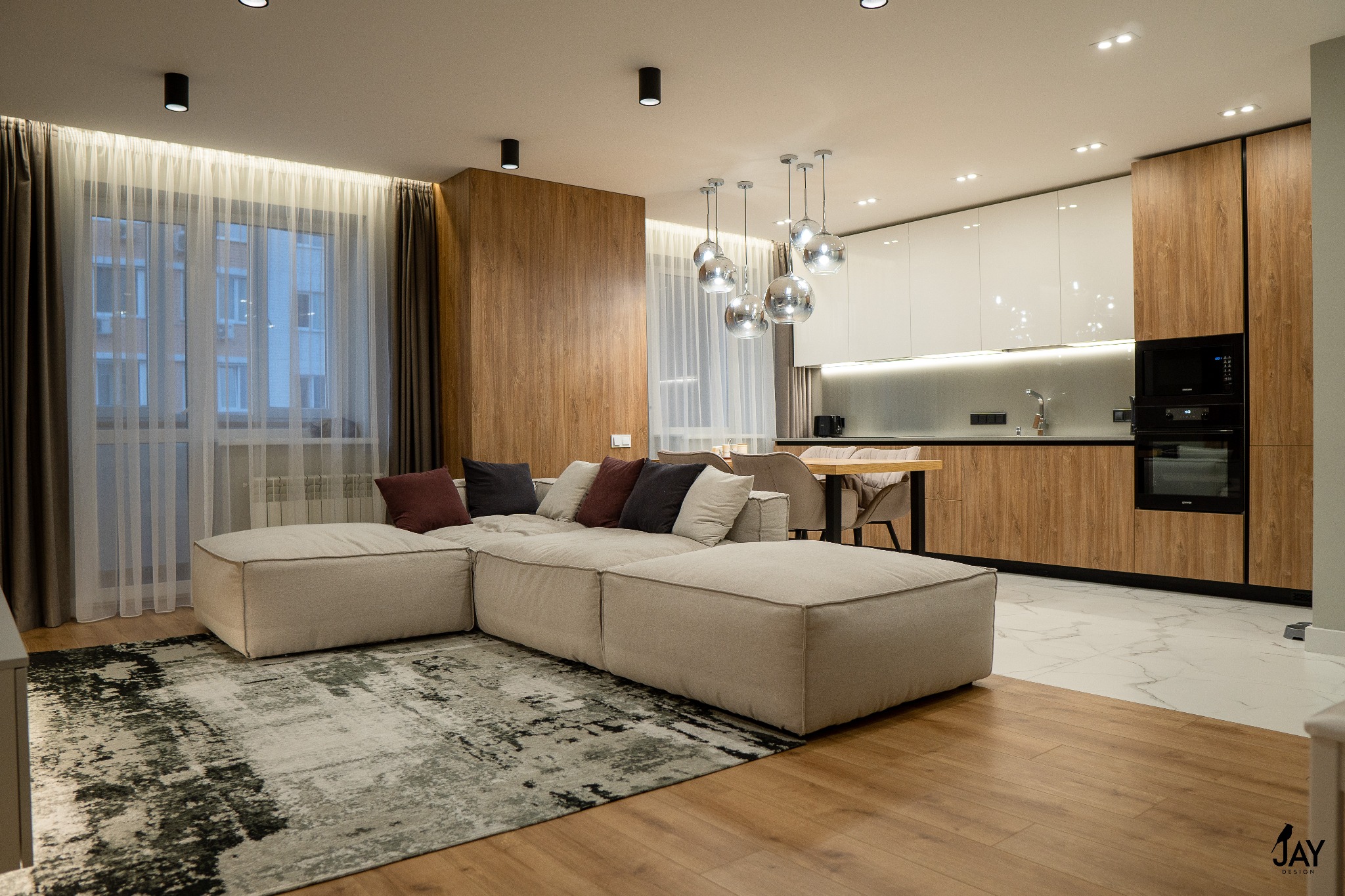 Nowoczesne wnętrze: beżowa modułowa sofa z poduszkami w odcieniach bordo i szarości na abstrakcyjnym dywanie, drewniana podłoga przechodzi w marmurową w strefie kuchennej z zabudową w kolorze...