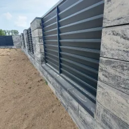 Nowoczesne ogrodzenie z paneli w kolorze grafitowym, osadzone na podmurówce z szarych, ciosanych bloków betonowych, widoczne z perspektywy ziemi, z fragmentem kolejnego przęsła w tle.