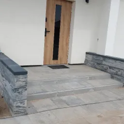 Wykończone wejście do domu z drewnianymi drzwiami z czarnym akcentem, schody i podest wyłożone jasnoszarą kostką brukową, niskie murki z szarej kostki po bokach, teren wokół jeszcze...