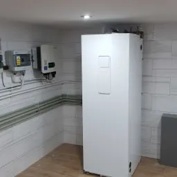 Biała pompa ciepła w pomieszczeniu z panelami ściennymi imitującymi cegłę i podłogą z płytek drewnopodobnych, widoczna skrzynka elektryczna i inwerter na ścianie.