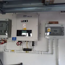 Instalacja fotowoltaiczna wewnątrz budynku: inwerter SOFAR z naklejką z flagą Japonii i tabliczką PV, dwie rozdzielnice Noark, białe rurki ochronne przewodów, widoczne okablowanie.