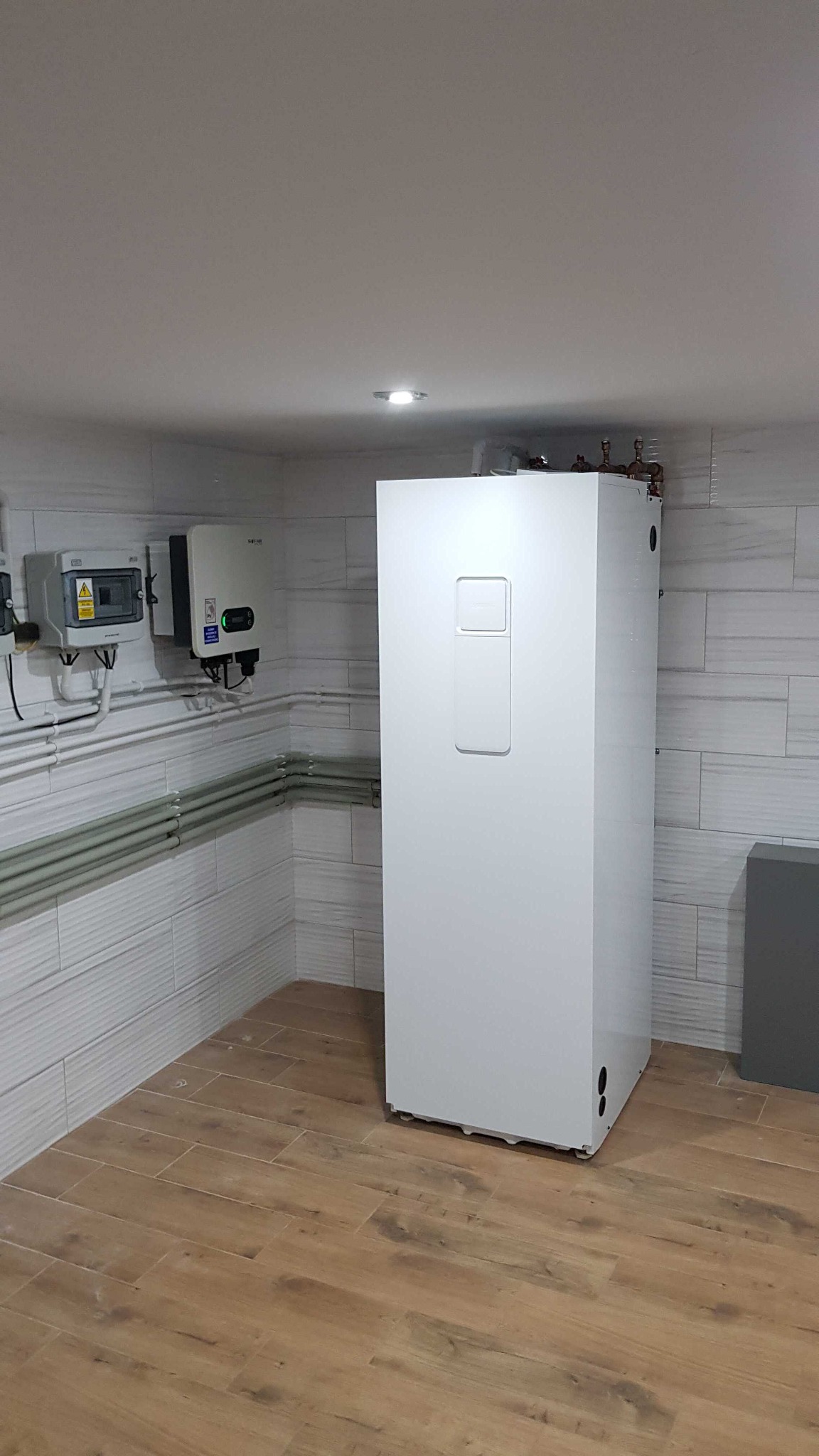 Biała pompa ciepła w pomieszczeniu z panelami ściennymi imitującymi cegłę i podłogą z płytek drewnopodobnych, widoczna skrzynka elektryczna i inwerter na ścianie.