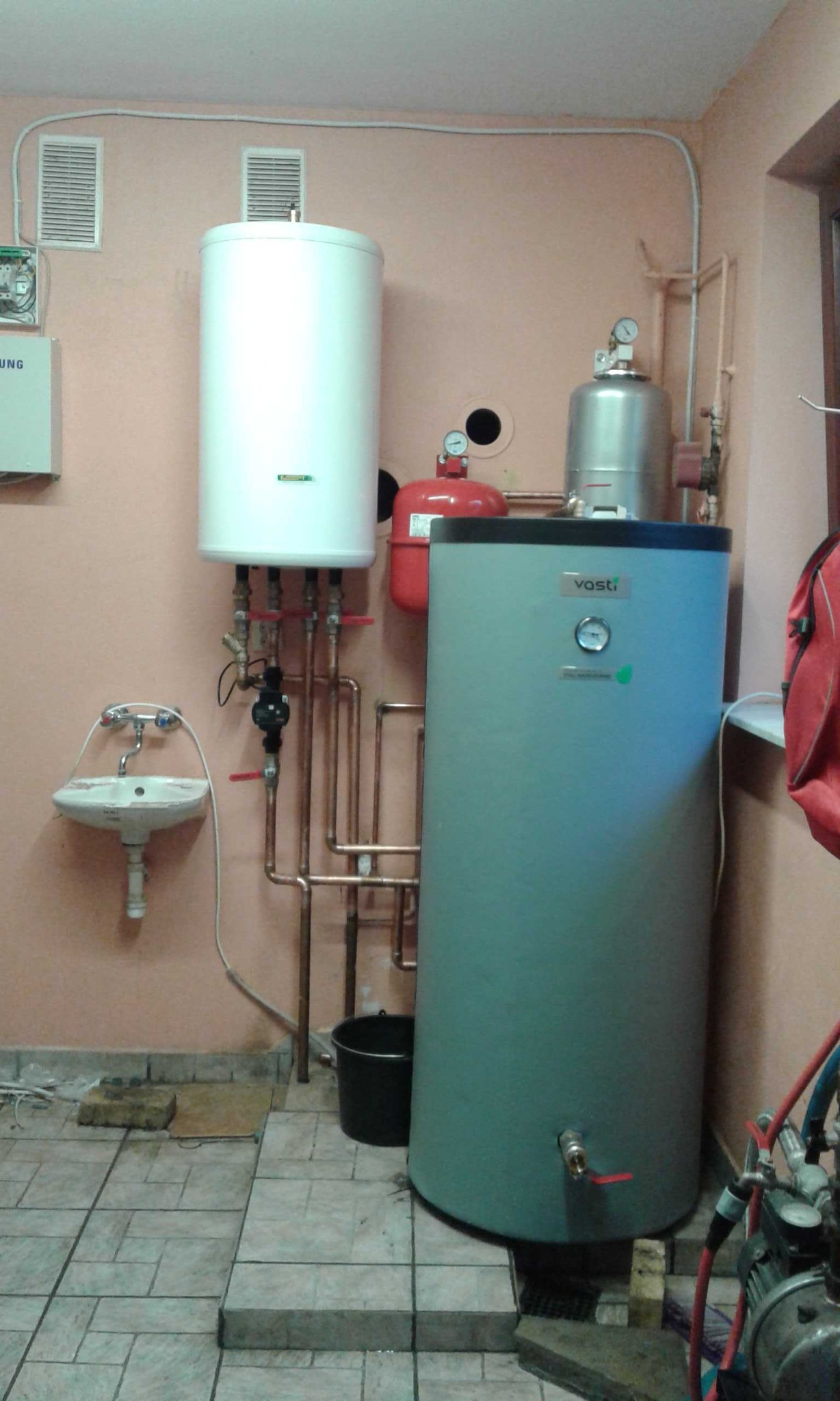 Instalacja grzewcza z dwoma zbiornikami, białym i szarym, oraz małą umywalką na ścianie w pomieszczeniu z kafelkami podłogowymi.