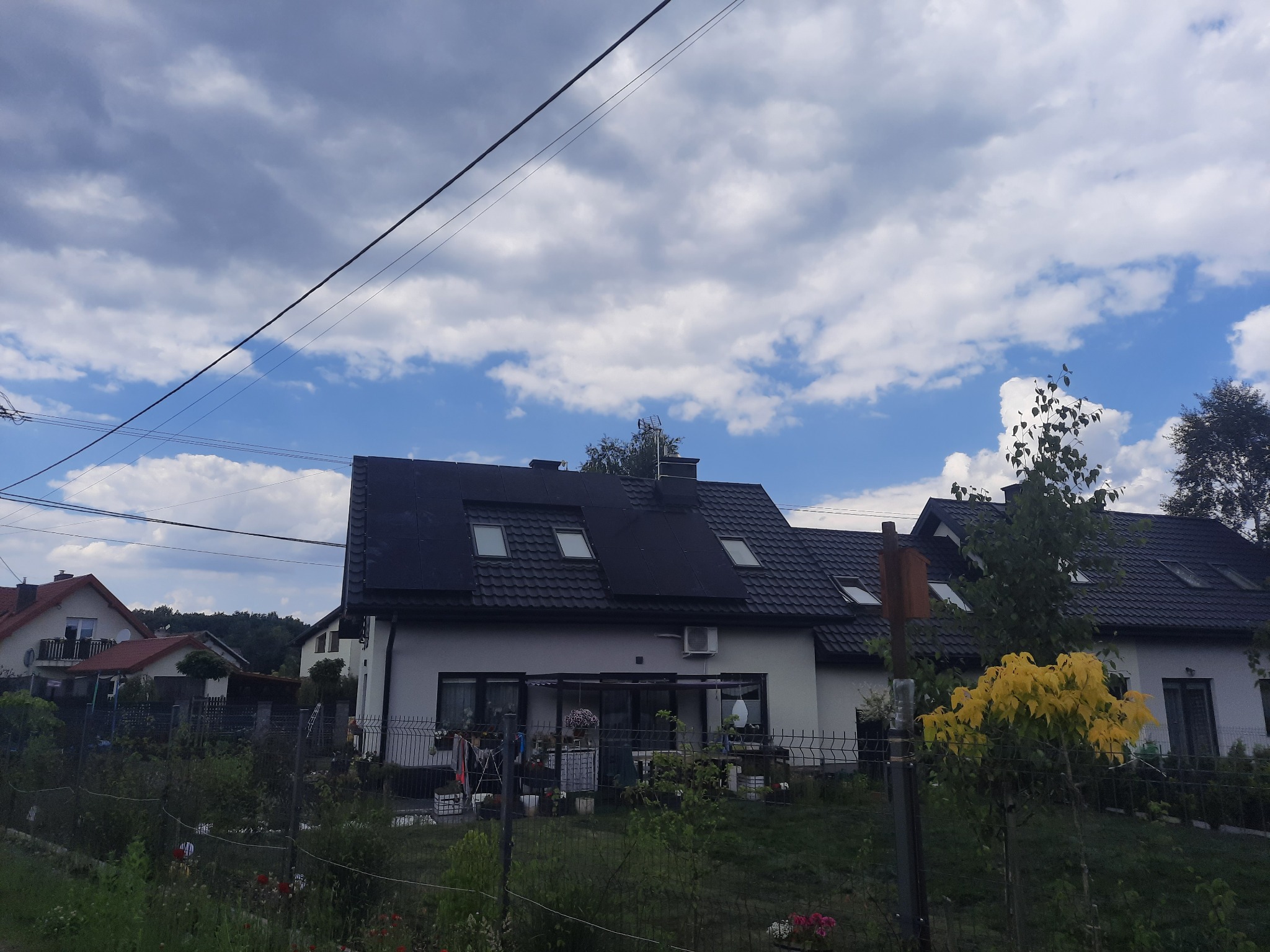 Dom jednorodzinny z panelami fotowoltaicznymi na dachu, widok zza ogrodzenia, pochmurne niebo.