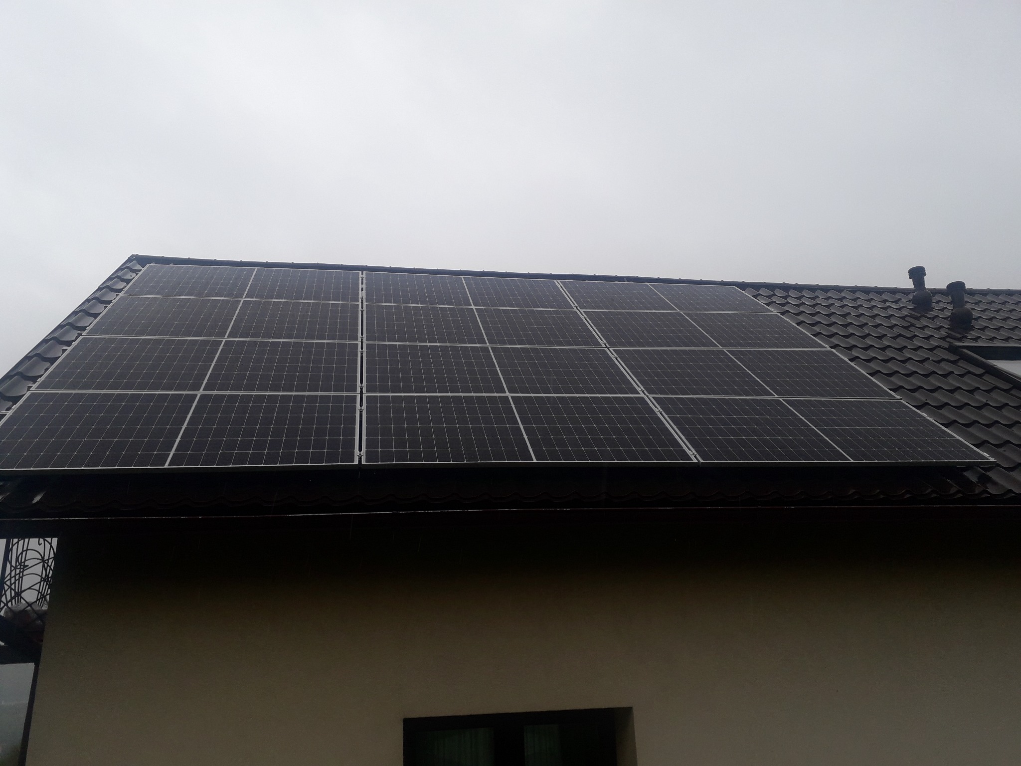 Panele słoneczne zamontowane na ciemnym, spadzistym dachu domu jednorodzinnego, widoczne kominy wentylacyjne, pochmurne niebo.