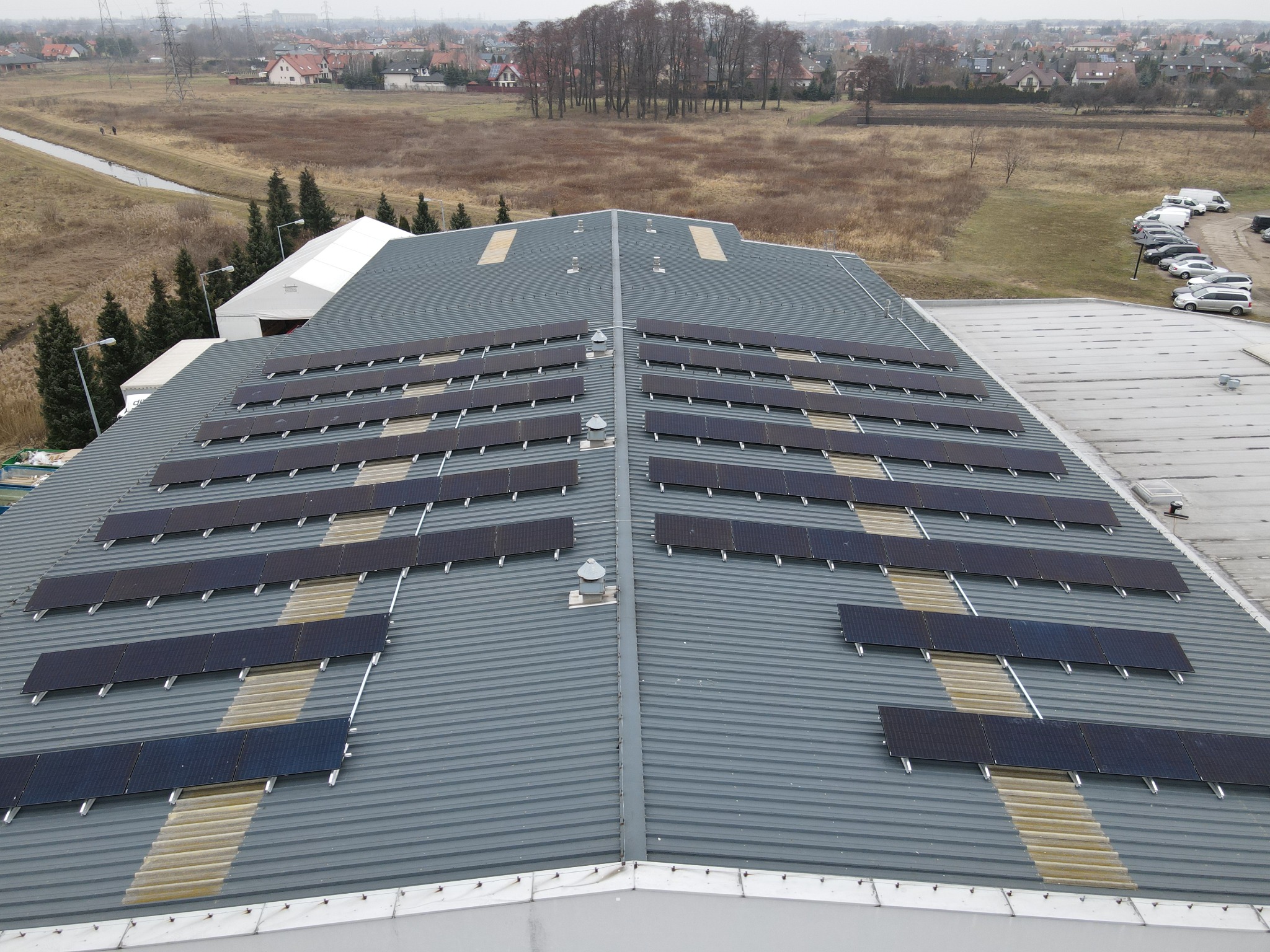 Rząd paneli słonecznych na szarym, metalowym dachu budynku przemysłowego, widok z góry, w tle drzewa i zabudowania.