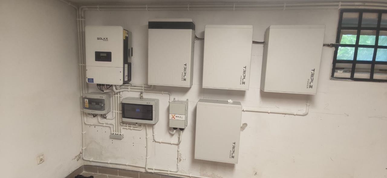 Instalacja domowa: inwerter Solax, liczniki i magazyny energii Triple Power zamontowane na ścianie w piwnicy, widoczne rury instalacyjne.