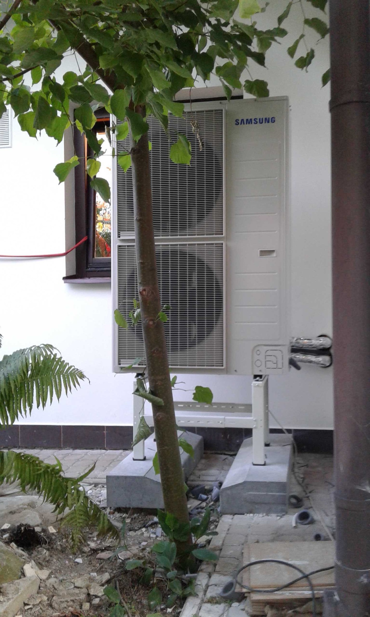 Zewnętrzna jednostka klimatyzacji Samsung na betonowym fundamencie, częściowo zasłonięta przez małe drzewo liściaste, widok z poziomu gruntu.