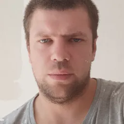 Portret mężczyzny z krótką brodą i plamami farby na twarzy i koszulce, stojącego na tle ściany w trakcie prac wykończeniowych.