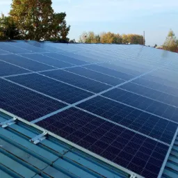 Panele słoneczne zamontowane na metalowym dachu z widocznymi elementami mocującymi, w tle drzewa i budynki w słoneczny dzień.