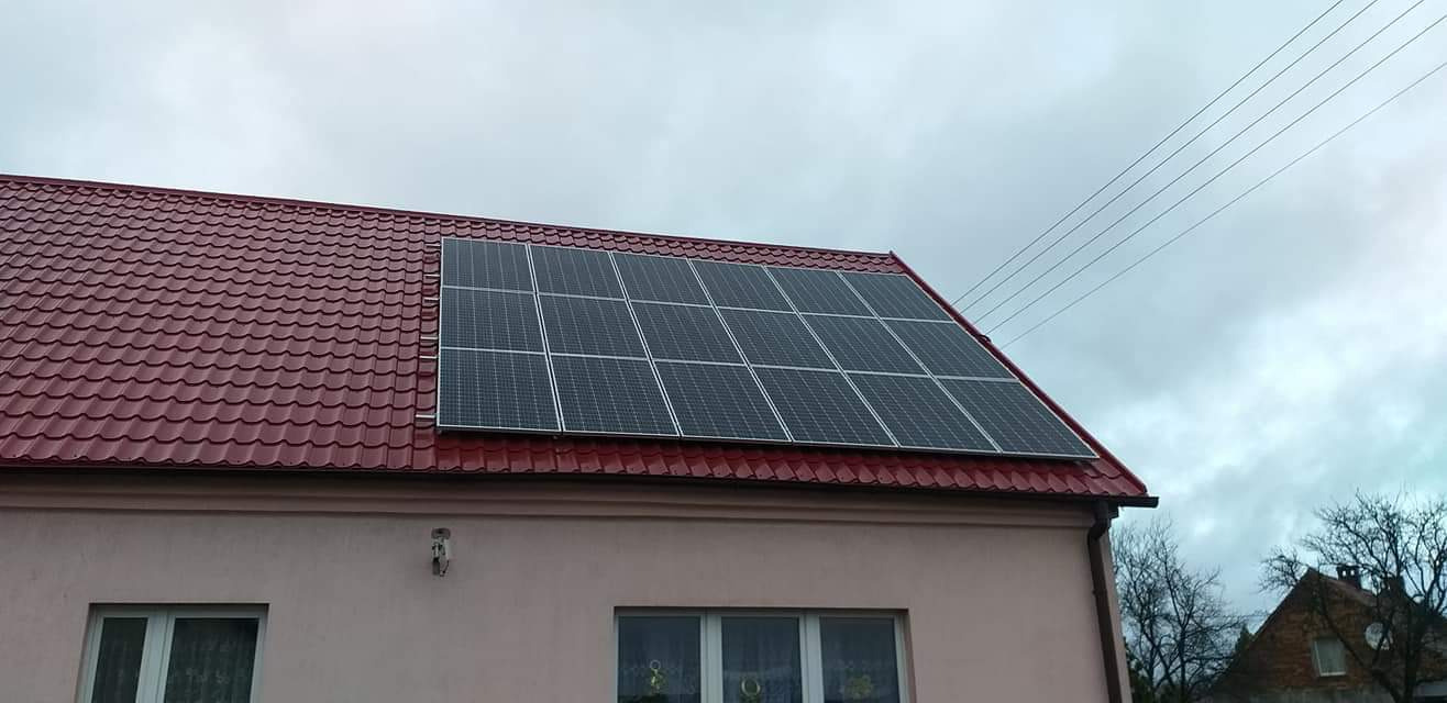 Panele fotowoltaiczne zamontowane na dachu domu z czerwoną dachówką, widoczne okna budynku i pochmurne niebo.