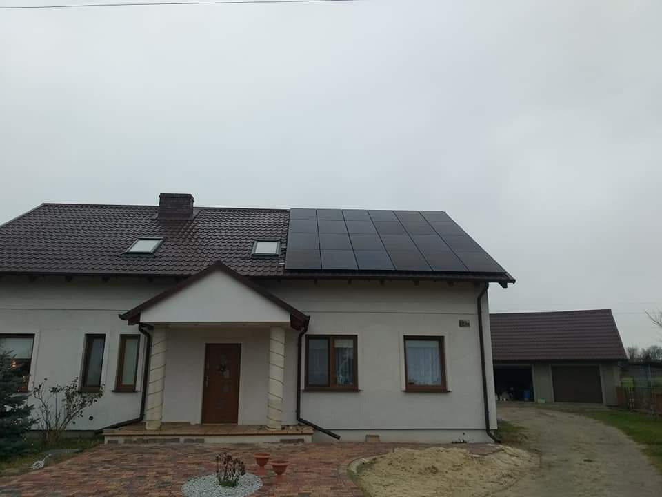 Dom jednorodzinny z brązowym dachem, na którym zamontowano czarne panele fotowoltaiczne. Widoczny komin, okna dachowe i podjazd z kostki brukowej.