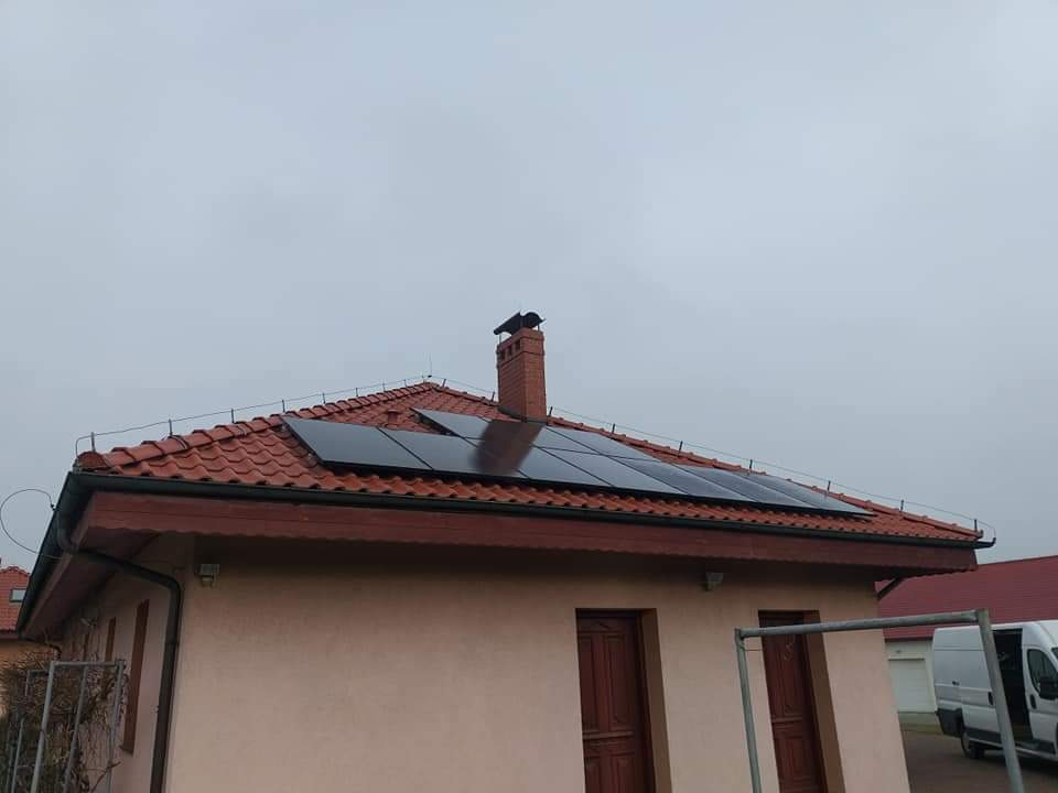 Panele słoneczne na dachu domu z czerwoną dachówką, komin z ptakiem na szczycie, pochmurne niebo.