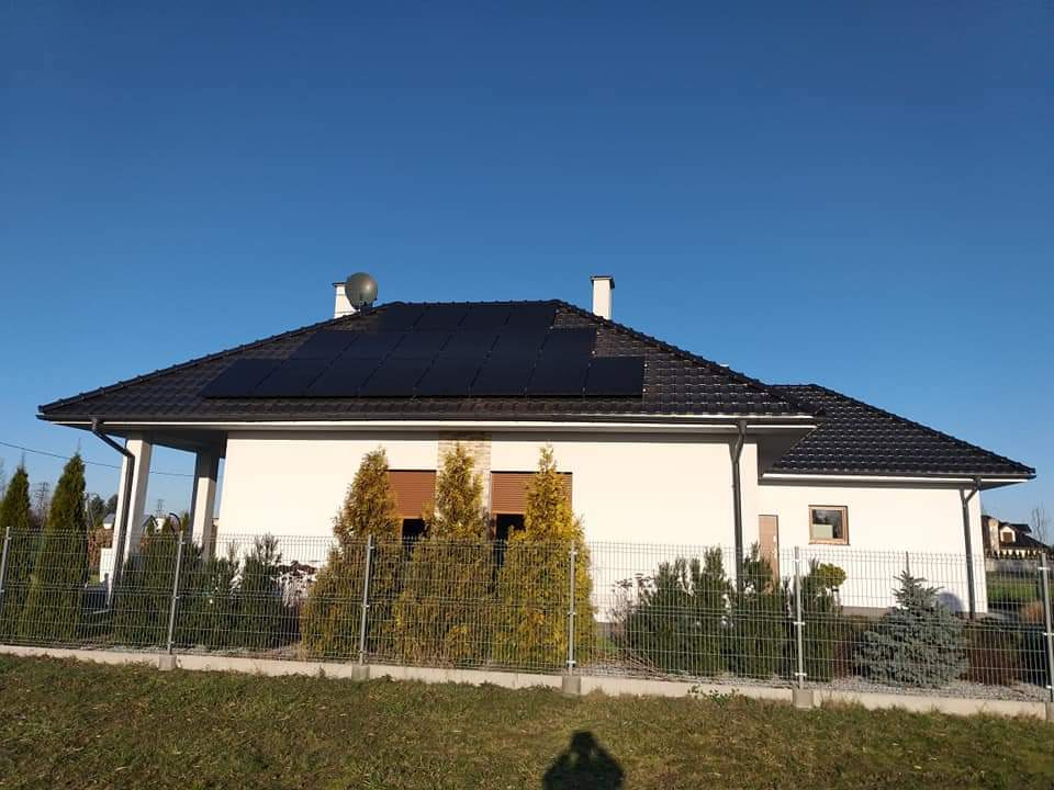 Biały dom jednorodzinny z ciemną dachówką i panelami słonecznymi na dachu, widok z poziomu gruntu, ogrodzony metalową siatką i ozdobnymi iglakami.