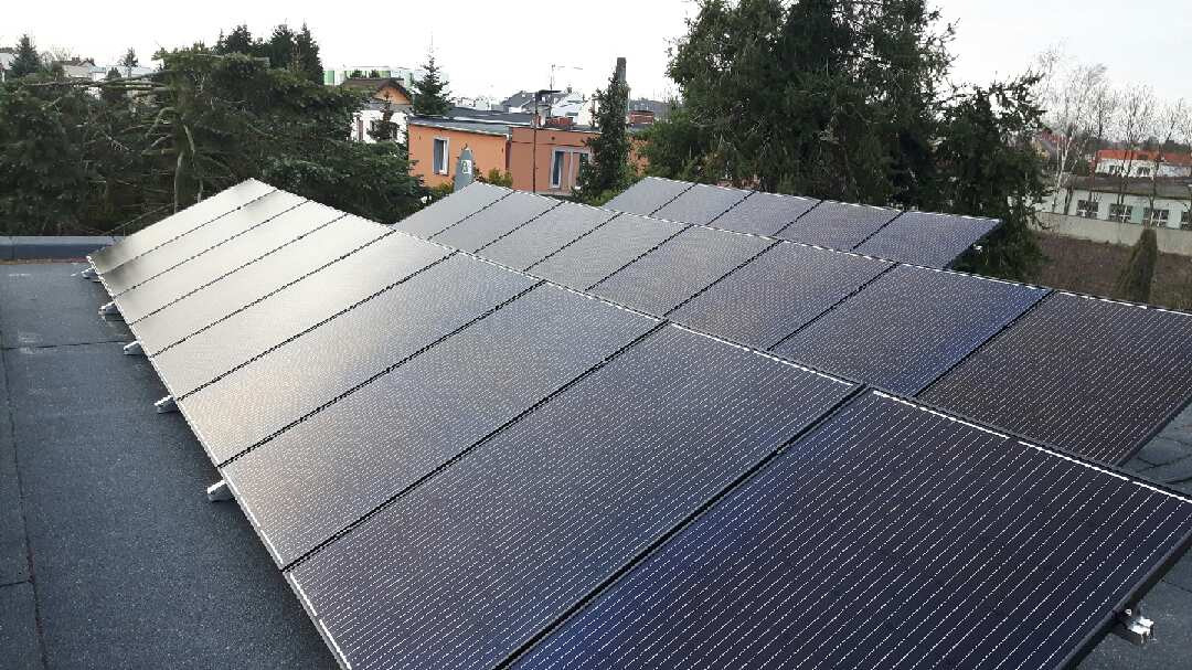 Rząd paneli słonecznych z widoczną strukturą montażową na płaskim dachu, w tle drzewa i zabudowa mieszkalna.