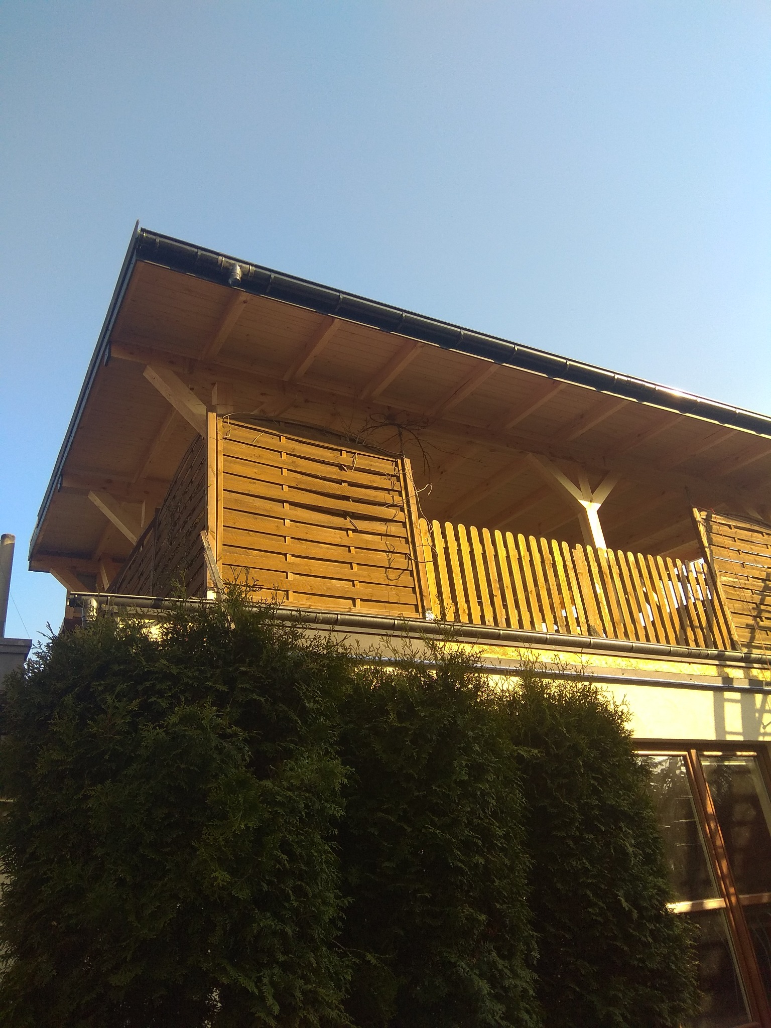 Widok z dołu na drewnianą konstrukcję dachu z rynnami, drewnianą balustradą balkonu i zielonymi krzewami na pierwszym planie w słoneczny dzień.