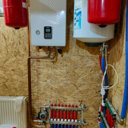 Sergii Cholavyn - Centrala grzewcza Navien, dwa zbiorniki wyrównawcze Stout w kolorze czerwonym, rozdzielacz z zaworami termostatycznymi i rurami w kolorze niebieskim i czerwonym na ścianie z płyty OSB.